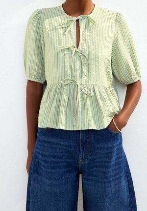 Personne portant un chemisier rayé vert clair avec trois liens à l'avant et des manches courtes bouffantes, assorti à un jean bleu foncé à jambes larges.
