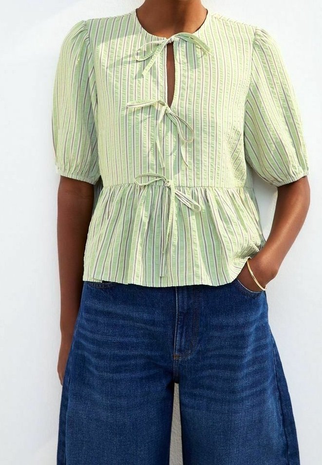 Personne portant un chemisier rayé vert clair avec trois liens à l'avant et des manches courtes bouffantes, assorti à un jean bleu foncé à jambes larges.