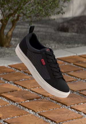 Zwartgrijze veterschoen met witte zool en rode "HUGO" labels, hangend boven houten vierkante tegels en grind buiten.