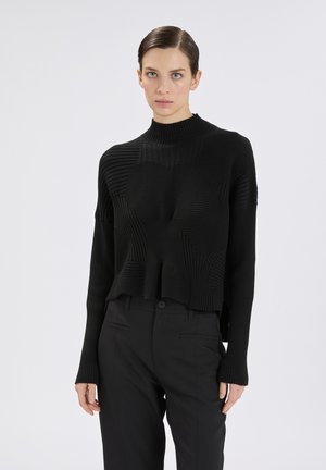 Donna che indossa un maglione dolcevita nero a coste e pantaloni neri a vita alta, in piedi contro uno sfondo grigio chiaro uniforme.