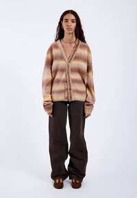 Hnědý a béžový pruhovaný kardigan s měkkou texturou, s výstřihem do V, zapínáním na knoflíky a dlouhými rukávy, v kombinaci s tmavými kalhotami.