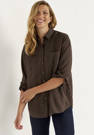Smilende kvinde med langt blond hår iført en mørk brun skjorte med knapper og optrukne ærmer samt mørkeblå jeans.