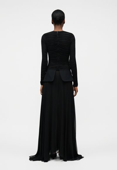 Žena s tmavou pletí a vlasy staženými do drdolu, oblečená v černém nátělníku s řasením na rukávech a volné černé dlouhé sukni, otočená zády na bílém pozadí.