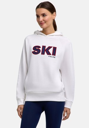 Vrouw draagt een witte hoodie met "SKI" in grote letters en "FALKE" eronder, gecombineerd met donkerblauwe broek, staand tegen een effen achtergrond.