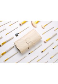 Beige lærmakeupbørste-rulle med en clasp, omgitt av pensler med gullfiner i ulike lengder og former på en hvit bakgrunn.
