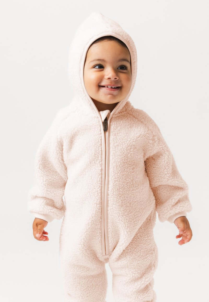 Nasmejan malček v svetlo rožnati kosmati kapuci s sprednjim zadrgo, stoji pred enobarvnim belim ozadjem.