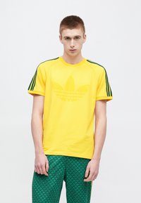 Żółta bawełniana koszulka z zielonymi paskami akcentującymi na rękawach oraz teksturowanym zielonym nadrukiem logo koniczyny na przodzie.