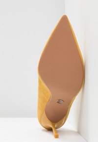 Chaussure jaune à talon haut et bout pointu vue par la semelle, appuyée contre un mur blanc sur une surface blanche.