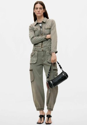 Femme portant une combinaison kaki avec plusieurs poches, tenant un sac à main cylindrique noir et portant des sandales noires sur un fond blanc uni.