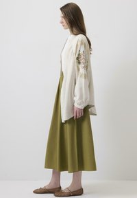 Cardigan lung de culoare bej deschis, cu mâneci brodate cu flori, asortat cu o fustă de culoare verde măslinie închisă și pantofi cu talpă din fibră naturală, maro.