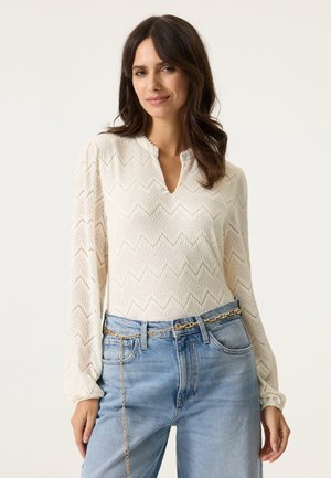 Witte blouse met lange mouwen en een chevronpatroon, voorzien van een v-hals en een textuurstof; gecombineerd met lichtblauwe jeans en een gouden kettingriem.