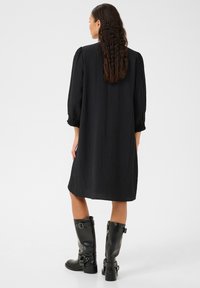 Robe noire en tissu texturé, manches bouffantes trois-quarts et coupe ample. Associée à des bottes en cuir noir hautes jusqu'aux genoux avec des boucles.