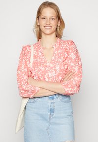 Blusa floral rosa com decote em camadas, mangas longas e fecho de botão. Combinada com uma saia de ganga azul clara. Tecido simples e leve.