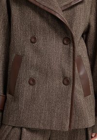 Giacca in tweed marrone con accenti in pelle marrone a contrasto, design doppiopetto e bottoni grandi, caratterizzata da un motivo a spina di pesce testurizzato.