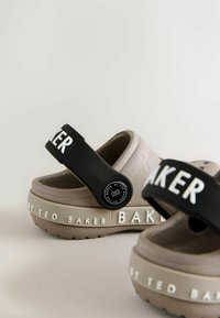 Beżowe obuwie z grubą podeszwą. Czarna, regulowana klamra z białym napisem. Teksturowana górna i dolna obudowa z detalami marki.