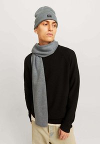 Beanie grigio a maglia e sciarpa abbinata, indossati con un maglione nero a trama. La sciarpa cade morbida attorno al collo, mettendo in risalto il suo materiale soffice.