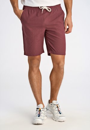 Bordeauxfärgade shorts med en texturerad yta, utrustade med resårmidja och dragsko. Matchas med ljusgrå sportskor.