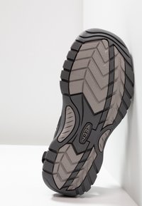 Black textured shoe sole featuring deep grooves and a pronounced tread pattern. Logo visible on the side, with a smooth grey midsole.