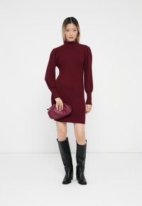 Knit, burgundy dress with a high neck and puffed long sleeves, paired with a small, dark red bag and black knee-high boots.
