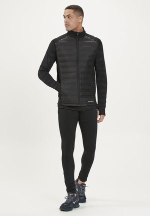 Zwarte pufferjack met een rits aan de voorkant, textuur op de zwarte mouwen en aansluitende zwarte leggings. Heeft een hoge kraag en ongespecificeerde branding.