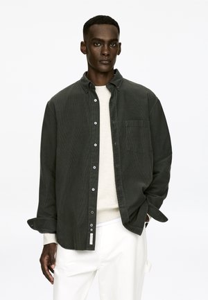 Dunkelgrünes Cordhemd mit Knöpfen, langen Ärmeln und einer Brusttasche. Übereinander getragen mit einem hellen Pullover, kombiniert mit weißen Hosen.