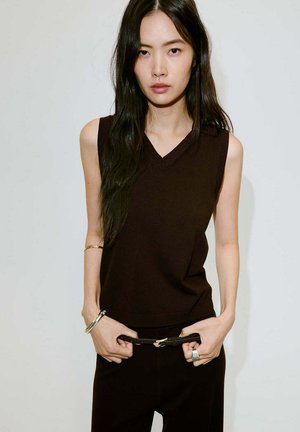 Jeune femme aux longs cheveux noirs portant un haut marron sans manches et un pantalon noir, ajustant une ceinture noire, sur un fond uni.