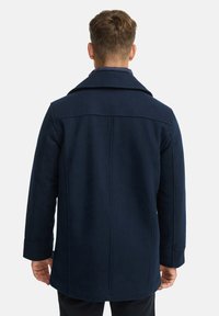 Mørkeblå uldsfrakke med en struktureret krave, lige snit og synlige sømdetaljer. Har lange ærmer og et mellemlangt design.