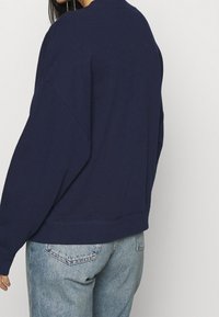 Námořnicky modrá mikina s volnějším střihem, spadlými rameny a žebrovanými manžetami a lemem. Kombinována s džínami v světlém odstínu.