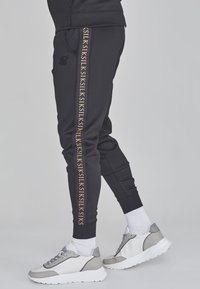 Schwarze Jogginghose mit einem goldenen Logo-Streifen an der Seite, aus weichem Material, kombiniert mit weißen und grauen Sportschuhen. Weiße Knöchelsocken sind sichtbar.