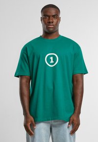 Camiseta de algodón verde con mangas cortas, que presenta un gran gráfico de círculo blanco y el número "1" en el centro. Corte relajado, diseño sencillo.