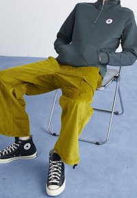Sudadera de forro polar verde oscuro con media cremallera, pantalones de pana amarillos y zapatillas deportivas negras de caña alta. Una silla y un suelo azul proporcionan contexto.