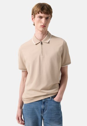 Młody mężczyzna w beżowej polówce z krótkim rękawem i suwakiem oraz niebieskich dżinsach, stojący z jedną ręką w kieszeni na jednolitym tle.