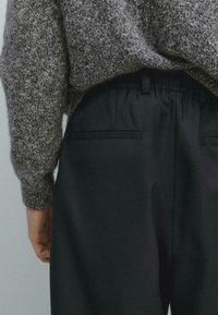 Pantalon noir avec une taille élastique, doté de deux poches arrière. Porté avec un pull en tricot texturé gris.