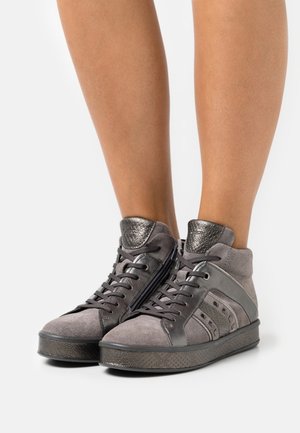 Beine in grauen High-Top-Sneakers mit Wildleder- und Metallic-Lederpaneelen, Schnüröffnung vorne und seitlichem Reißverschluss auf weißem Hintergrund.