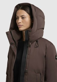 Brown puffer jacket with a hood, featuring black accents, a zippered front, and snap buttons. Soft texture with a quilted design.