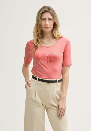 T-shirt rose corail à manches courtes avec un col rond et texte imprimé "soleil" dans différentes polices. Associé à un pantalon taille haute beige.