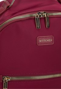 Burgundy tygryggsäck med guldfärgade dragkedjor, med en framträdande "Wittchen" logotypapplikation och minimala sömnadsdetaljer.