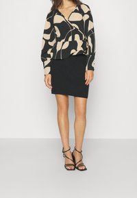 Schwarze und beige gemusterte Wickelbluse mit langen Ärmeln, kombiniert mit einem figurbetonten schwarzen Ripp-Minikleid und schwarzen Sandalen mit Absatz und Riemchen.