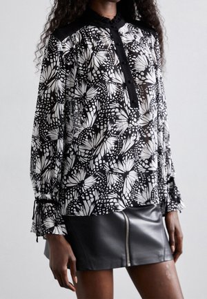 Femme portant un chemisier noir et blanc à motif papillon avec des poignets à volants et une mini jupe en cuir noir avec une fermeture éclair avant.