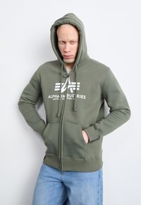 Olivovo zelená mikina na zips s predným logom, kapucňou so šnúrkou a klokankovými vreckami, kombinovaná so svetlomodrými džínami. Jednoduchý a neformálny dizajn.