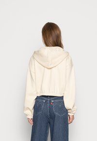 Osoba v krémové zkrácené mikině s kapucí dolů a modrých širokých džínách s proužky, viděná zezadu na jednobarevném bílém pozadí.