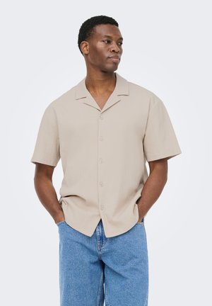 Homme portant une chemise beige à manches courtes boutonnée et un jean bleu, debout les mains dans les poches, regardant sur le côté sur un fond uni.