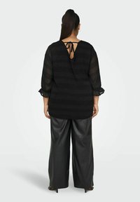 Blusa nera in tessuto trasparente e testurizzato, con scollatura a V e dettaglio a fiocco sul retro. Abbinata a pantaloni in pelle nera con vestibilità ampia.