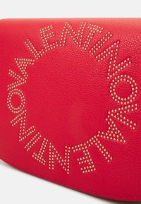 Röd läderväska med cirkulär design och nitar som stavar "Valentino." Texturerad yta med metalliska detaljer.