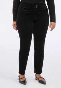 Persona vistiendo pantalones negros ajustados, top de manga larga negro con destellos y zapatos planos negros puntiagudos con correas en el tobillo, de pie sobre un fondo blanco.