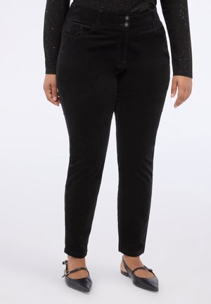 Persona vistiendo pantalones negros ajustados, top de manga larga negro con destellos y zapatos planos negros puntiagudos con correas en el tobillo, de pie sobre un fondo blanco.