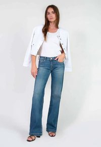 Veste en denim blanc portée sur un t-shirt blanc, associée à un jean flare bleu clair. Mannequin portant des talons noirs, accessoires minimalistes, sur un fond neutre.