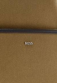 Καφέ υφασμάτινη υφή με λεπτό μαύρο δερμάτινο περίγραμμα και ασημένιο μεταλλικό λογότυπο "BOSS" κεντραρισμένο κάτω από το περίγραμμα.