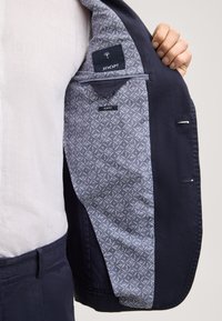 Homme tenant un blazer bleu marine ouvert montrant la doublure intérieure grise à motifs avec les étiquettes "JOOP!" et "SLIM FIT".
