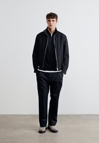 Νεαρός άνδρας που στέκεται σε εσωτερικό χώρο, φορώντας μαύρο μπουφάν, μαύρο παντελόνι, λευκή φανέλα και ανοιχτό γκρι παπούτσια, με φόντο απλό λευκό τοίχο.